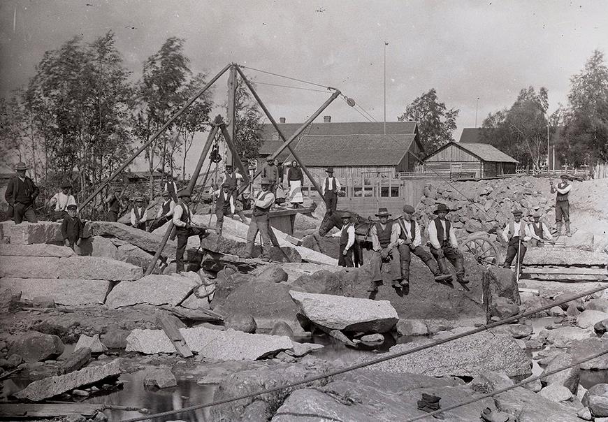 Här på Viktors foto så ser vi byggarna i full fart med byggandet av den bastanta dammen i Pärusfors. Den unga pojken till vänster i bilden är Viktors son Rurik. Kvisgården i bakgrunden. En tillfällig damm hade byggt högre upp i ån som ledde vattnet till den södra fåran, så att byggarna kunde bygga i fred. 