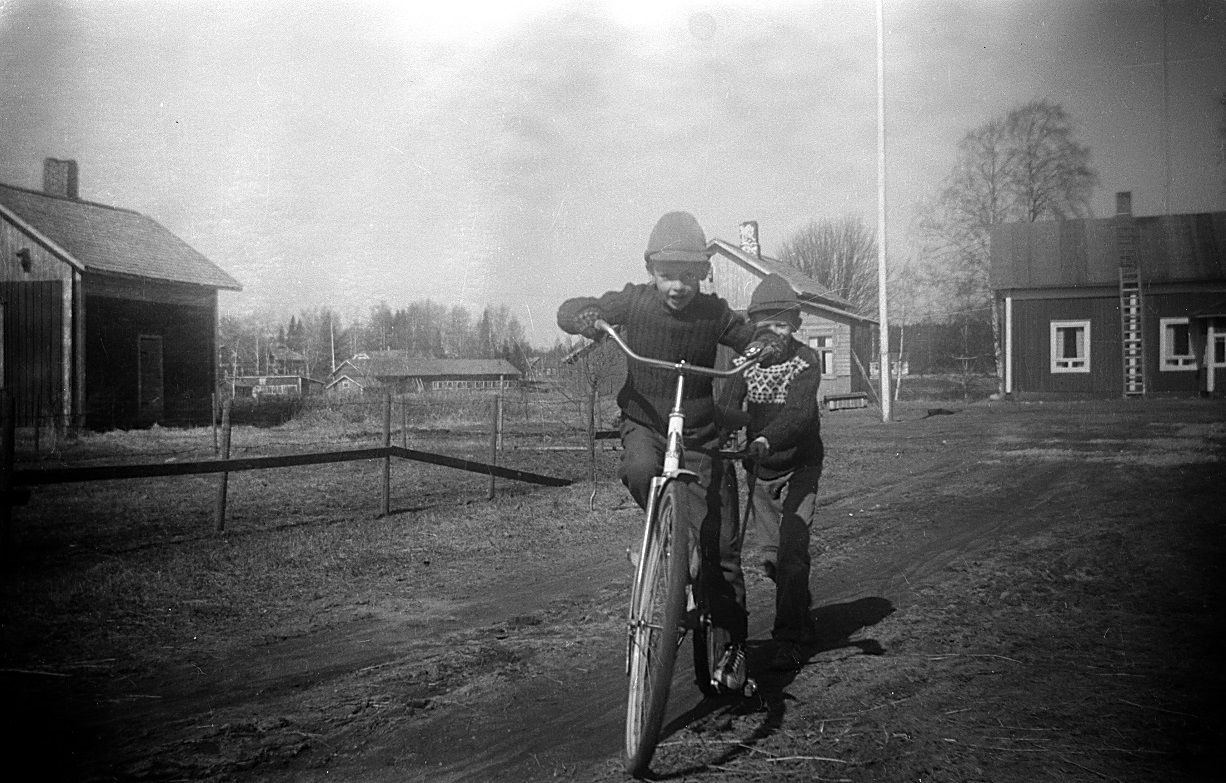 Lasse på cykeln och John springer efter och håller i. På andra sidan ån syns uthuset hos Åke Klemets. Folkskolan skymtar också i vegetationen.