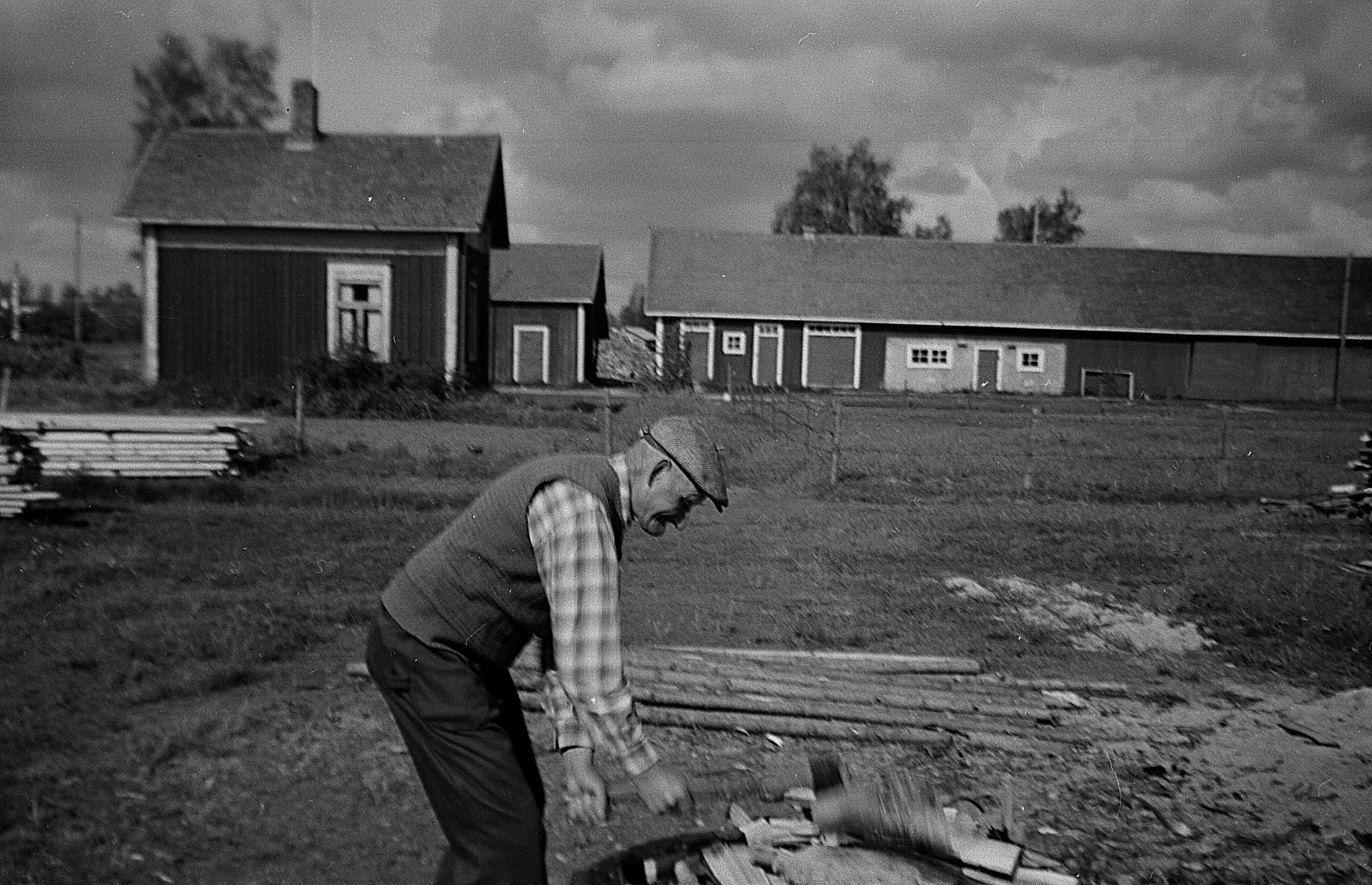 Den här bilden av Frans Agnäs klyvande ved åt sin syster Ida Backlund på vedbacken är tagen i början på 1950-talet. Byggnaden till vänster användes som bastu och längre bak syns uthusen som inrymde stall, fähus och röckhus. 