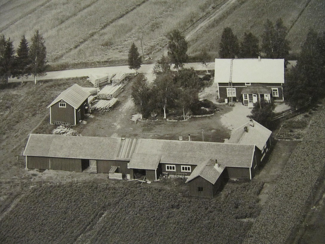 Den här flygbilden är från år 1961 och Myllyniemis gård uppe till höger. Den högra ingången gick till snickeriet och den vänstra till Johannes och Sylvias bostad. Snickarvirket finns upplagrat nära landsvägen. Åkervägen på andra sidan Åbackvägen leder till Oskars gård, som revs år 1961.