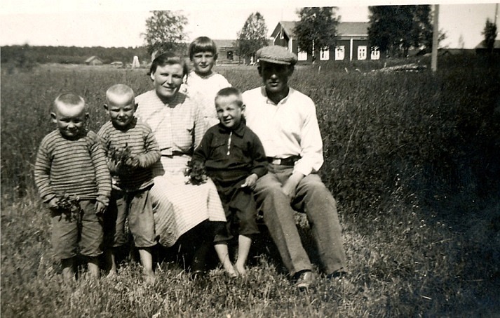 Här sitter hela Santamäkis familj i gräset på Åbackan och i bakgrunden syns ”Åsinas-Viktoras” alltså Viktor Rosenbacks gård som i slutet på 20-talet övertogs av Anselm Viklund då han återvände från Amerika. 
