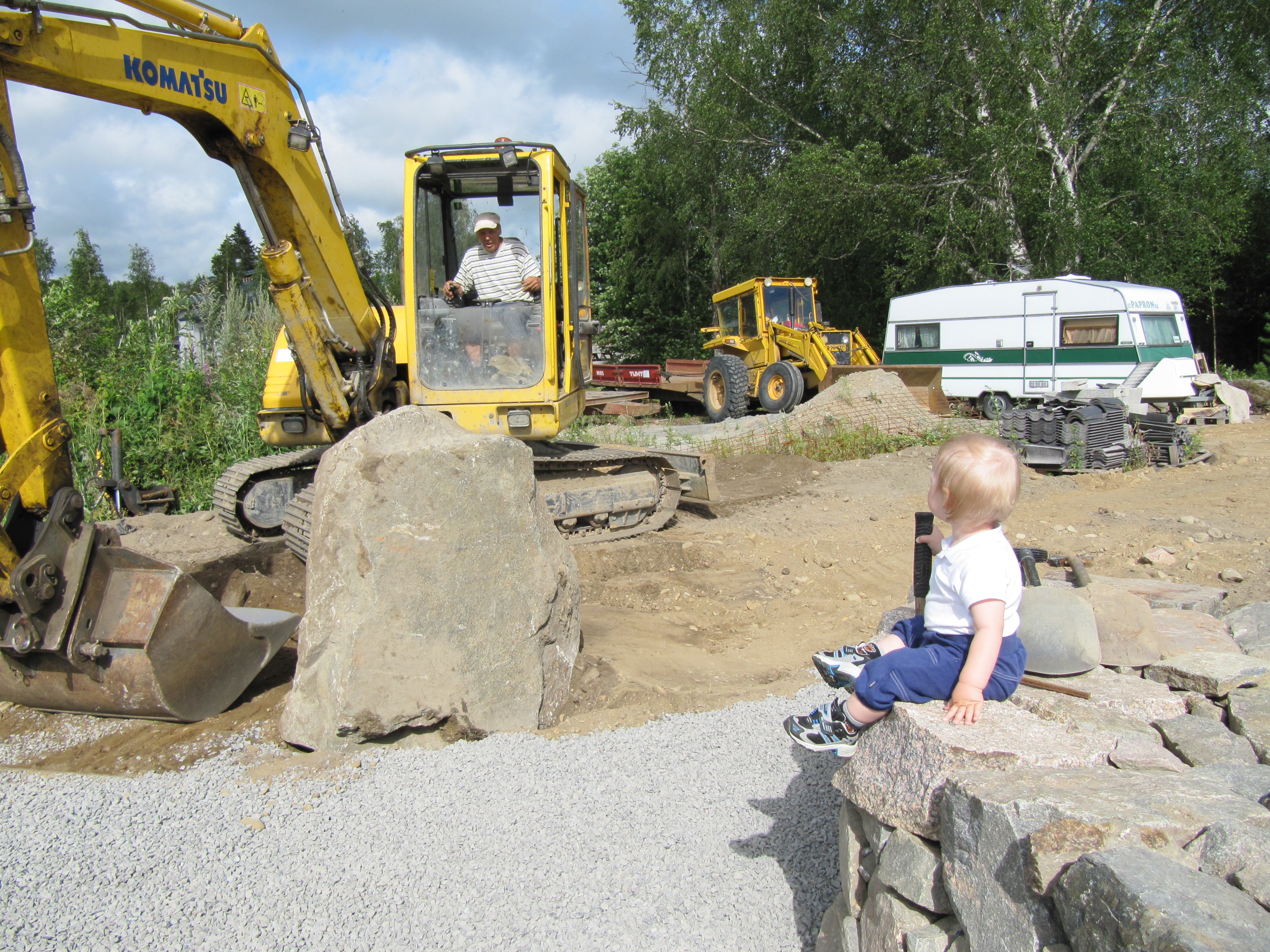 Den lilla pojken på stenmuren ser beundrande på då Jukka flyttar stora stenblock med grävmaskinen.