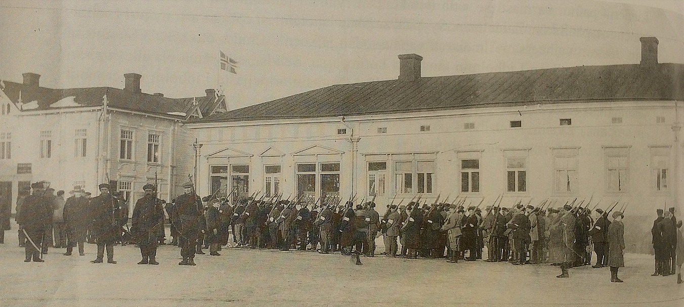 Den 31 januari 1918 var en stor dag i Kristinestads historia, då de ryska trupperna och deras bundsförvanter rödgardisterna avväpnades på morgonen efter en kort strid. Här ser vi skyddskårerna uppställda på torget den 31 januari. Flaggan i bakgrunden på Töttermans gård är den tillfälliga röda flagga med gult kors med blåvita kanter som användes en kort tid efter Finlands självständighetsförklaring.