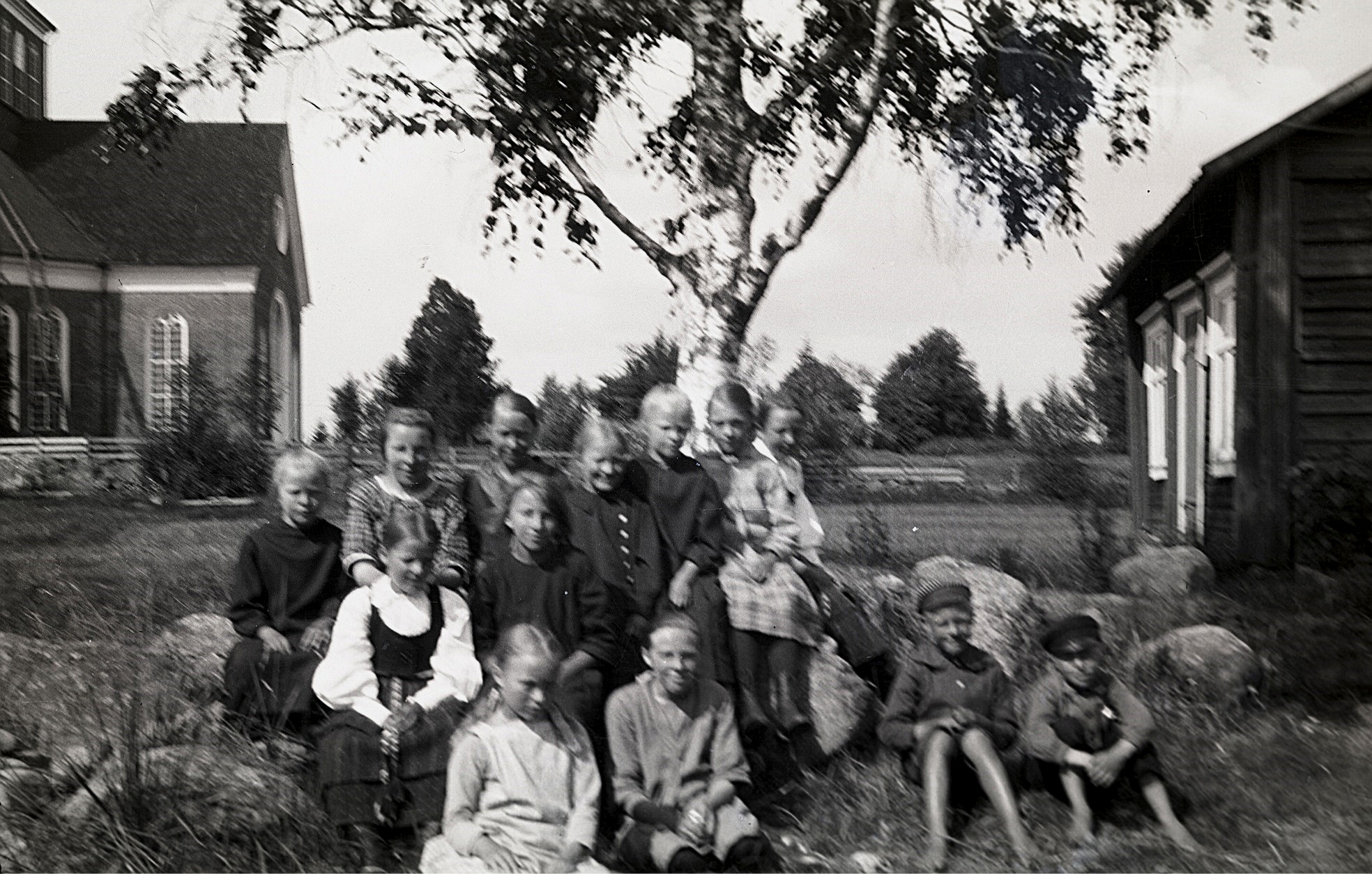 Barn på skolans gårdsplan med skolbastun till höger och kyrkan till vänster. Flickan längst fram till vänster är Ellen Asplund och bredvid henne sitter Helmi Ekman. Flickan i folkdräkt är Elin Rosenback och där bak finns bland annat Tyra Ingves, Märta Skogman, Anna Rosenback, Elin Gullmes, Svea Ådjers och Hilda Svedberg. Den av pojkarna som sitter till vänster är Karl Ådjers.