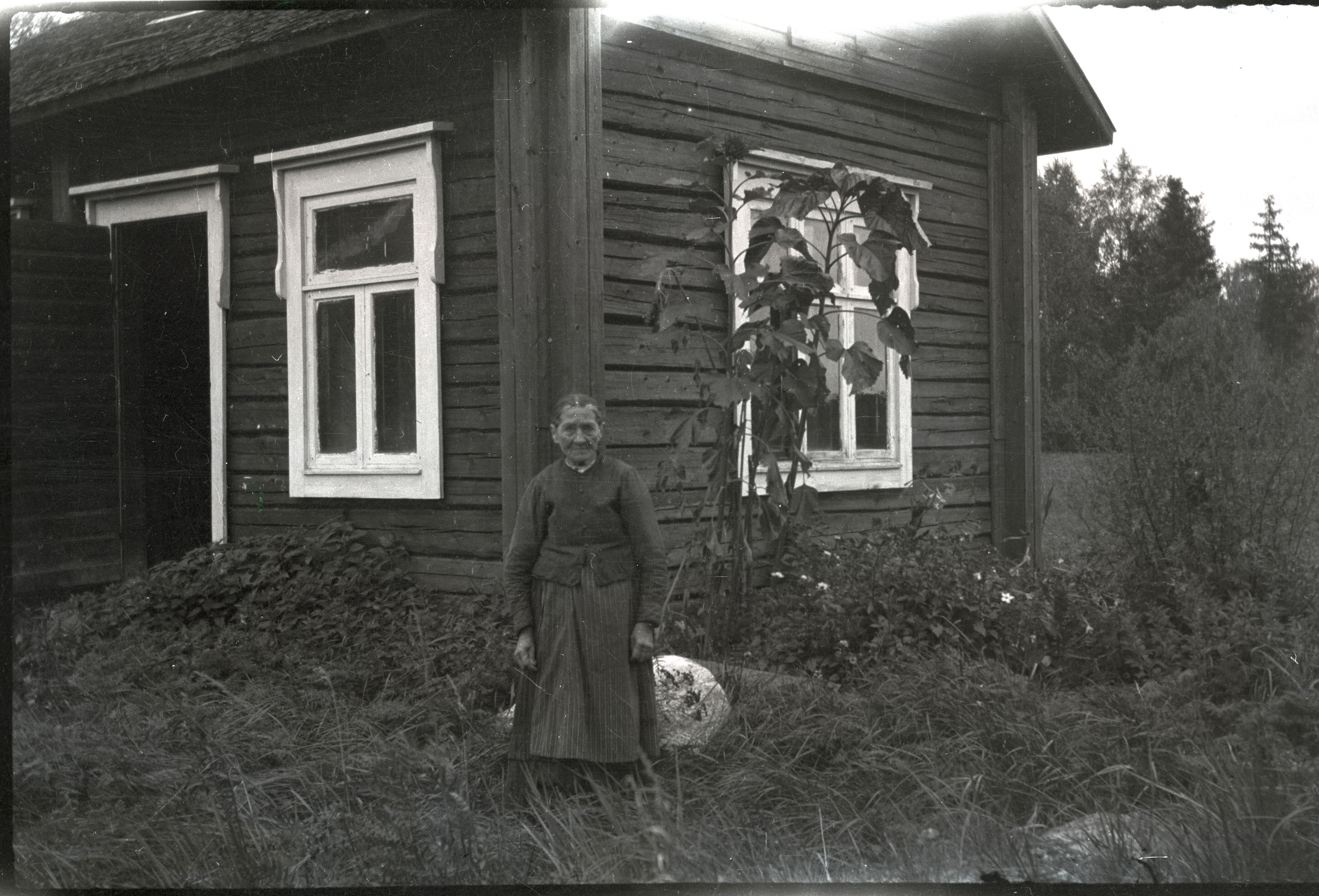 "Skol-Gret" var lärarinna Basts trotjänarinna som tvättade, städade, kokade, sprang ärenden, hämtade mjölk och handlade. En liten späd kvinna med långa armar, här fotograferad utanför skolbastun och tvättstugan. Själv bodde hon 200 meter söderut från skolan i en stuga som sedan Sandra Granqvist övertog och efter henne Eva, Elsa och Anni Vesterlund.