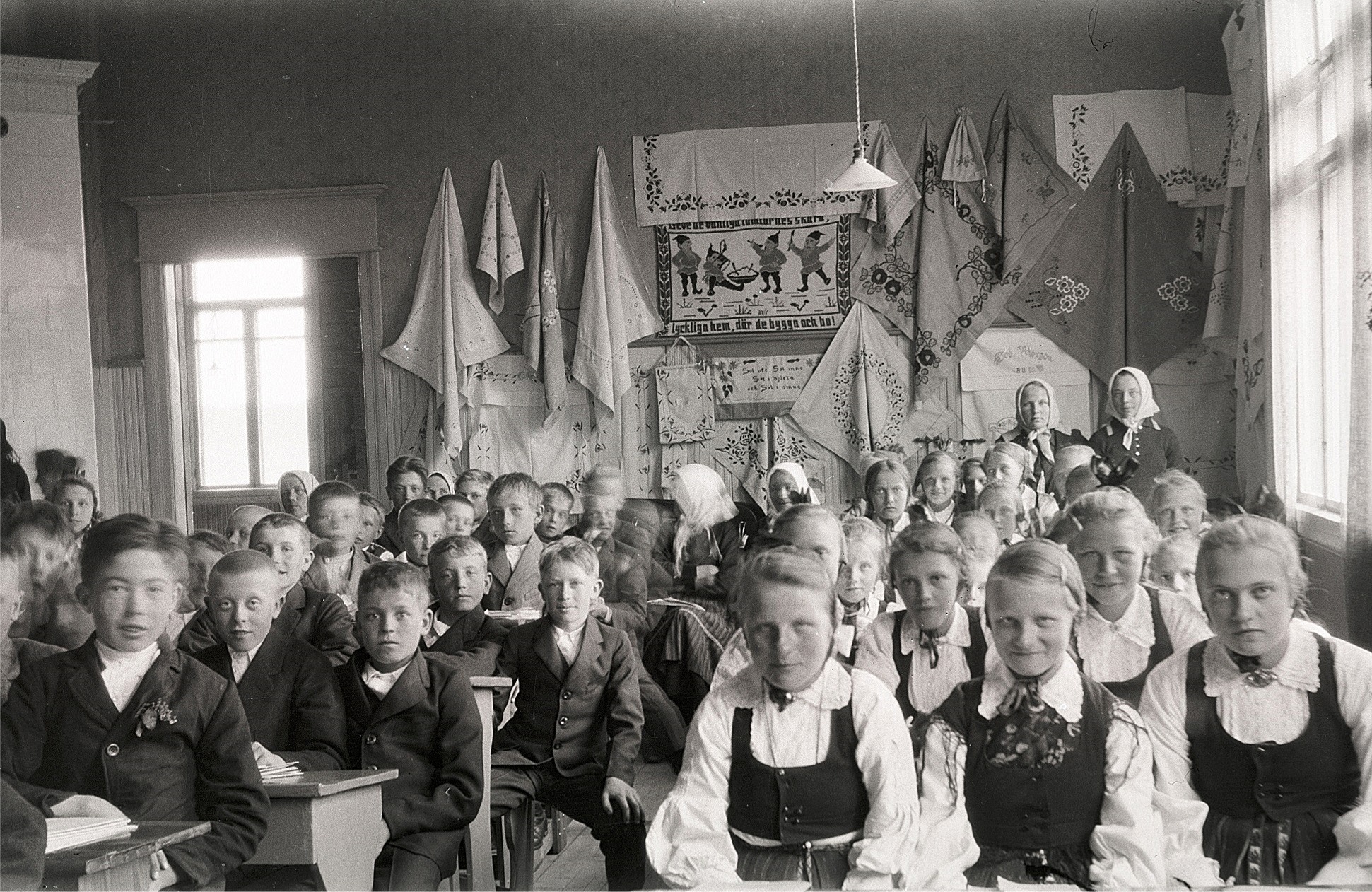 Foto från skolavslutningen 1930, där flickornas handarbeten finns upphängda på väggen. Till vänster sitter Arvo Haavisto och Karl Malm. Flickorna är Märta Skogman, Svea Ådjers, Ellen Norrholm, Elin Mangs och Alice Björses. Pojken mitt i bild med handen på knät är Valter Björknäs och bakom honom en pojke med huvudet på sned och det är Erik Storkull, senare Stens, som sedan gjorde ett stort arbete med att identifiera alla dessa Selim Björses fotografier.