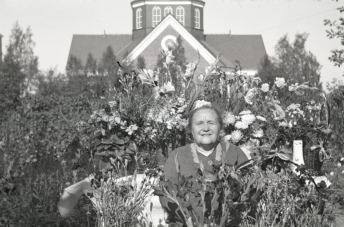 Lärarinnan Emmi Bast uppvaktades rikligt med blommor då hon fyllde 65 år och här sitter hon ute i trädgården utan lärarbostaden.