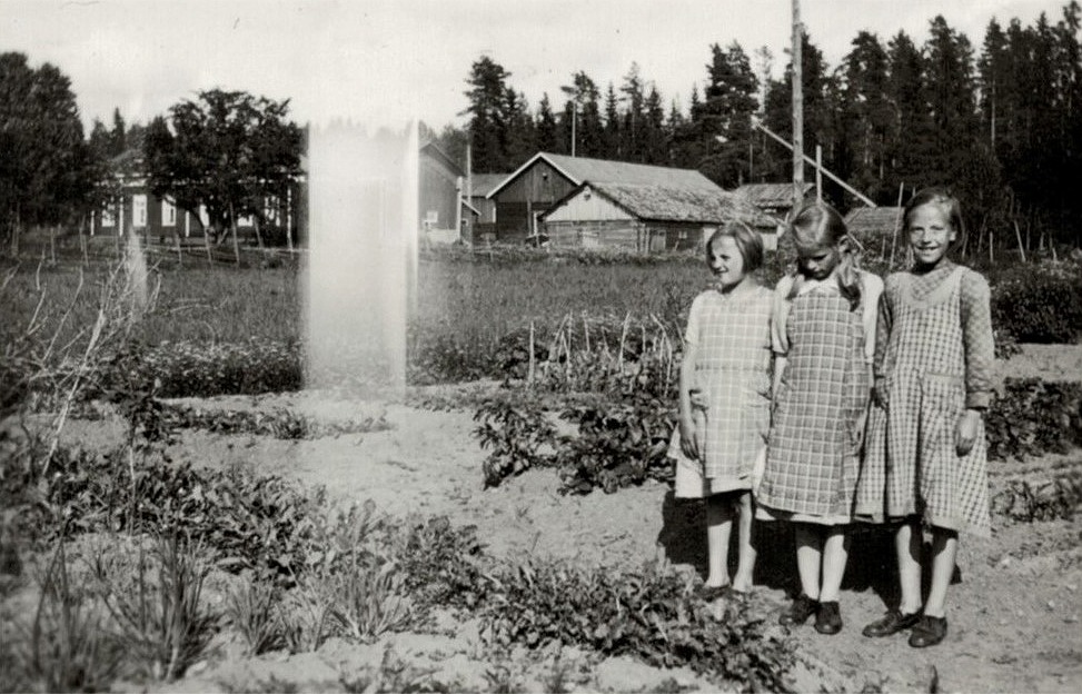 Tre korsbäckflickor står vid Sylvia Liljedahls (f.1928 senare Stenlunds) trädgård. Stenlunds ståtliga gård i bakgrunden.