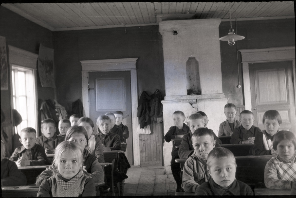 Så här satt eleverna i den gamla småbarnsskolan på Kladdbackan. Här är den sista årskursen 1932-33, sedan flyttade småskolan till södra folkskolans tillbyggnad. Dörrarna bak i salen gick in till lärarinnan Tekla Skrivars rum. Notera den speciella lampan i taket. I raden längst till vänster sitter Ingmar Nyqvist, Alfons Nyqvist, Gösta Gullmes, okänd. I följande rad mot höger sitter Rut Rosenback, Gunvor Juth, Ellen Haavisto och Elvi Lassfolk. Raden på andra sidan mittgången sitter Nils Mitts, Johan Hellman, Per Klockars, Uno Lassfolk, Birger Hellman. Längst till höger sitter Judit Hellman, Maj-Britt Granö, Gustav Hellman, okänd.