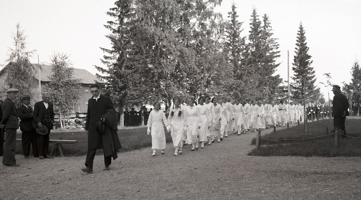 Över 100 konfirmander på väg in i kyrkan på midsommardagen 1936, flickorna i vita dräkter och pojkarna där bakom i mörka kostymer och vita handskar. Först i raden kommer systrarna Anna och Helga Englund från Dagsmark, sedan Ellen Wissander, Elin Holm, Hjördis Söderkvist, Gunni Björs och många till.