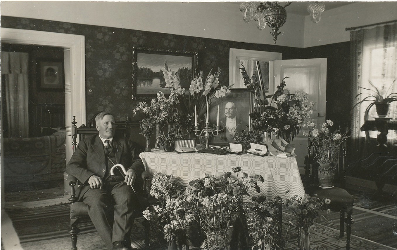 Här sitter Viktor Nylund bredvid bordet fyllt med fina blommor och presenter på sin 60-årsdag år 1933. På bordet står president Svinhufvuds porträtt.