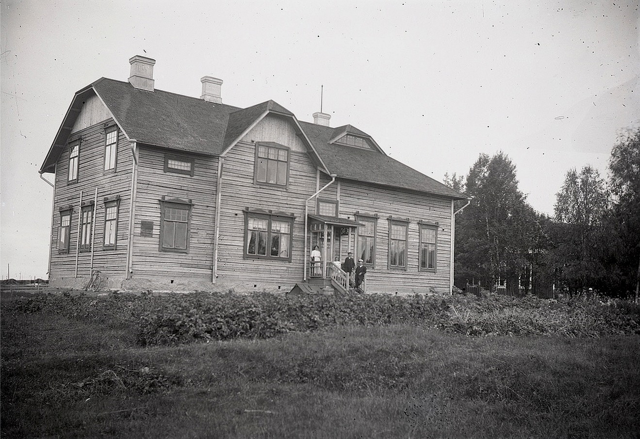 Här den splitternya skolan fotograferad år 1909, där några av lärare Wadströms barn stående på vindstrappan. Till höger skymtar den gamla skolan som revs följande år, då en del av virket kunde användas för byggandet av folkskolan i Perus. Foto Viktor Nylund.