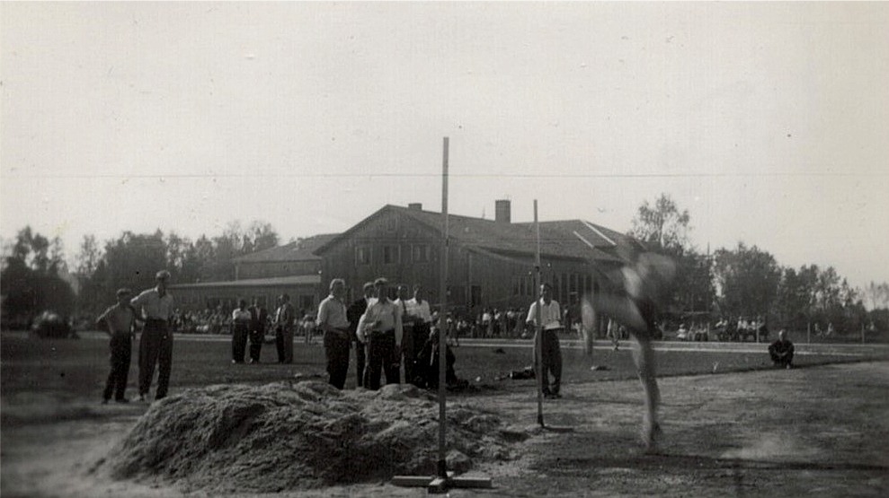 En höjdhoppare tar sats för att hoppa över ribban, för att sedan landa i spånhopen. I bakgrunden den ännu omålade ungdomslokalen. Fotot från invigningen av den nya sportplanen 21.8.1955.