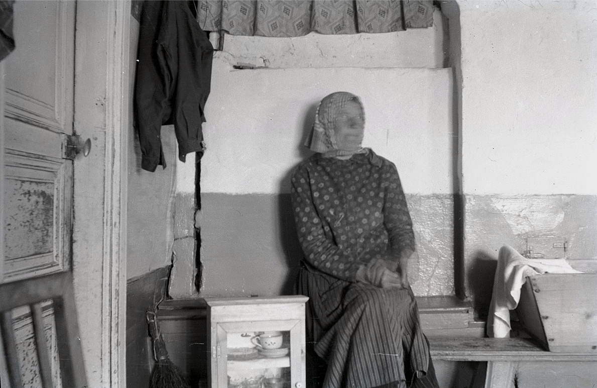 Gammal kvinna sitter på "spisbre" i sitt enkla hem.