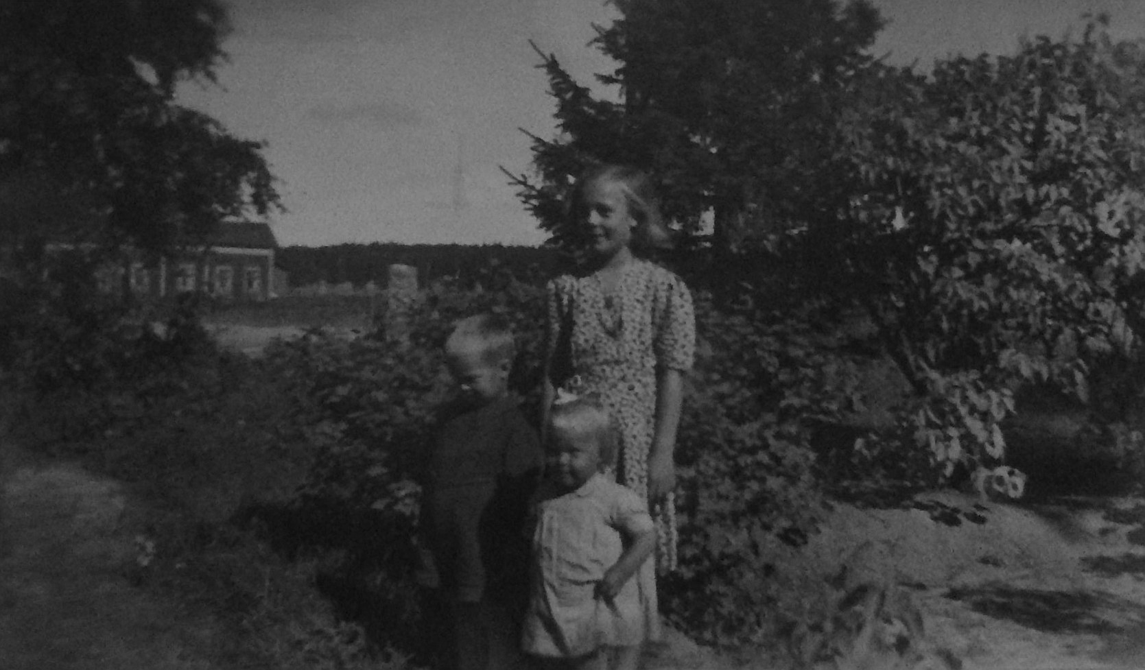 Syskonen Jarl, Annel och Gun-Lis Grans i trädgården. I bakgrunden syns "Roraas-Viktor" Rönnkvists gård.